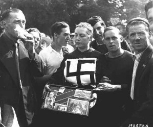 Une femme tondue, au milieu d'hommes, le 9 août 1944. Elle tient dans ses mains des photos et porte sur la poitrine une croix gammée.